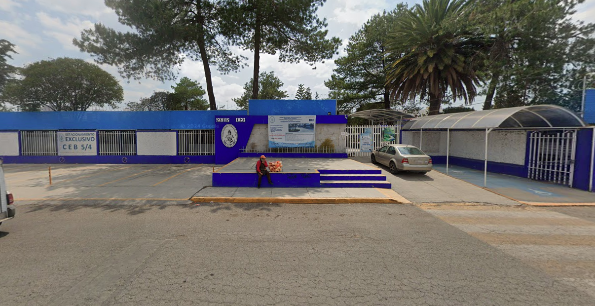 Fotografía en la que se muestra la fachada del plantel, conformada por muros y rejas de color blanco intercalados. Sobre uno de los muros, en letras azules, dice ce e be. A la izquierda de la foto hay un pasillo techado que lleva a la reja de acceso.